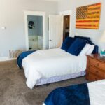 A bright bedroom with two beds, white bedding, and dark blue pillows. There is a wooden nightstand with a lamp, a dresser, and a U.S. flag artwork on the wall. A bathroom is visible through an open door.