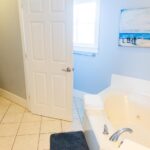 A bright bathroom with a large white bathtub, a toilet partially visible behind an open door, a teal laundry basket, and a painting of a dog and beach scene on the wall. Natural light comes through a window.