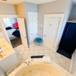 Bright bathroom with a large bathtub, two sinks, a mirror, a laundry basket, two white doors, a shower with a navy curtain, and wall signs. Bedroom with a red chair is visible through an open doorway.