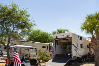 A golf cart with an American flag is parked next to large RVs under a clear blue sky, with one RV ramp open revealing equipment inside, surrounded by trees and palm trees.