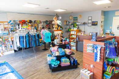 A beach-themed gift shop interior with racks of clothing, hats, towels, cups, and souvenirs. Shelves display various colorful items and accessories. The shop has wood flooring and light blue walls.