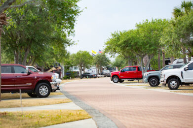 A paved road curves through an RV park lined with several parked pickup trucks and trees. Flags can be seen in the distance, and the scene appears calm, with greenery and RVs throughout the area.