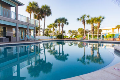 A curved outdoor swimming pool surrounded by lounge chairs and palm trees, with a modern building on the left and clear blue sky reflected in the calm water.