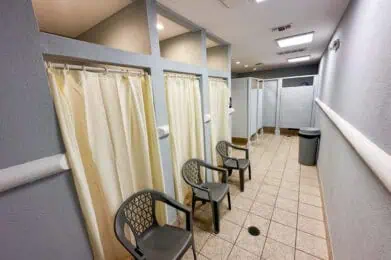 A clean locker room with tile floors, three shower stalls with cream-colored curtains, three black plastic chairs, a trash bin, and privacy partitions for toilets in the background. Bright overhead lights illuminate the space.