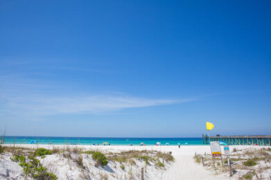 A sandy beach with clear blue water, colorful umbrellas, and a wooden pier. A yellow flag waves on a pole near the entrance, and green vegetation is scattered on the white sand under a bright, cloudless sky.
