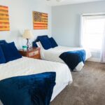 A bright bedroom with two double beds with white quilts and navy blue pillows and throws, a wooden nightstand with a lamp, a red armchair, a window with white curtains, and two American flag artworks on the wall.