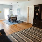 A bright, spacious living and dining area with wood floors, a striped rug, a navy couch with patterned pillows, a small dining table with four chairs, a TV cabinet, and large windows letting in natural light.