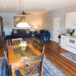 Open-plan living and dining area with a wooden table and chairs, sofas, a rug, white kitchen appliances, light-colored walls, and a ceiling fan. A vase with flowers sits on the dining table.