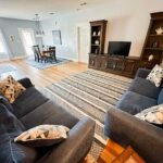 A spacious living room with two dark blue sofas, a striped area rug, a wooden TV stand with a flat-screen TV, and a dining area with a table and chairs in the background. Natural light fills the space.