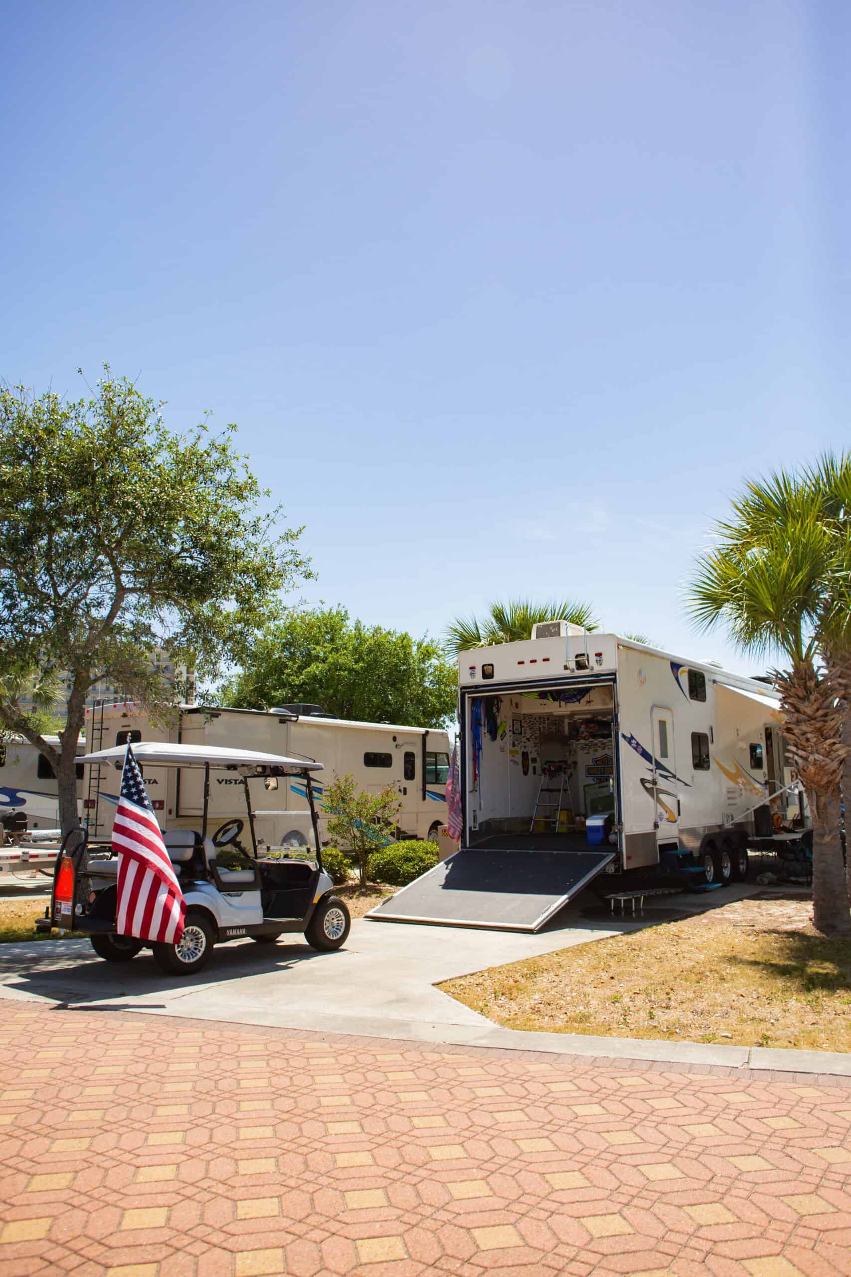 A golf cart with an American flag is parked near a large RV with its back ramp open in a sunny, paved camping area with trees and palm trees.