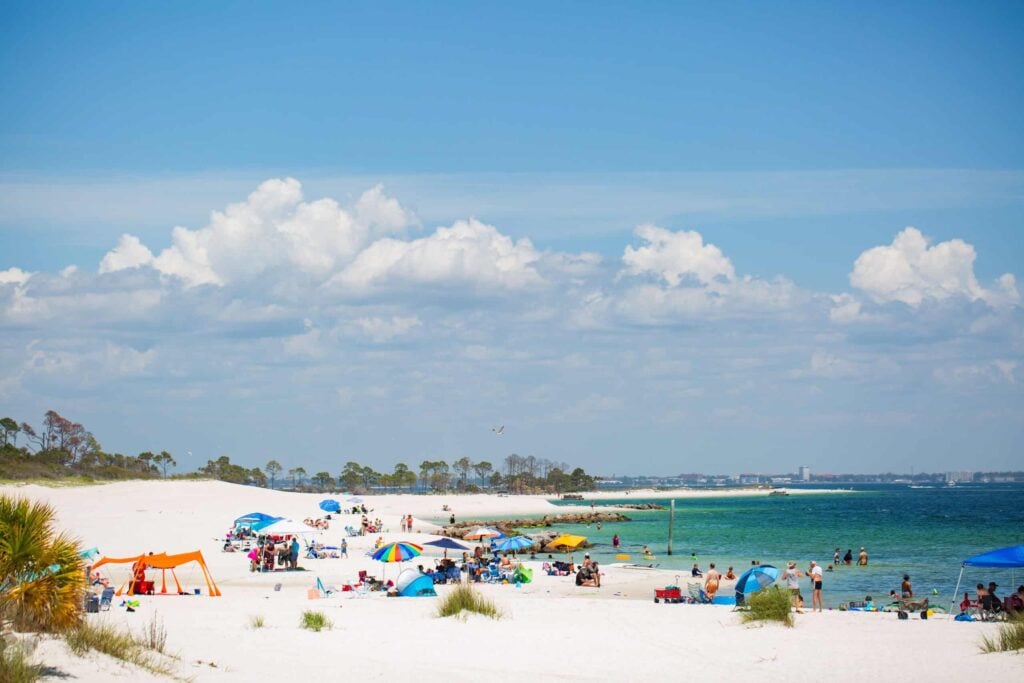 People relax on a sandy beach under colorful umbrellas near clear blue water, with scattered clouds in the sky and distant trees along the shoreline.