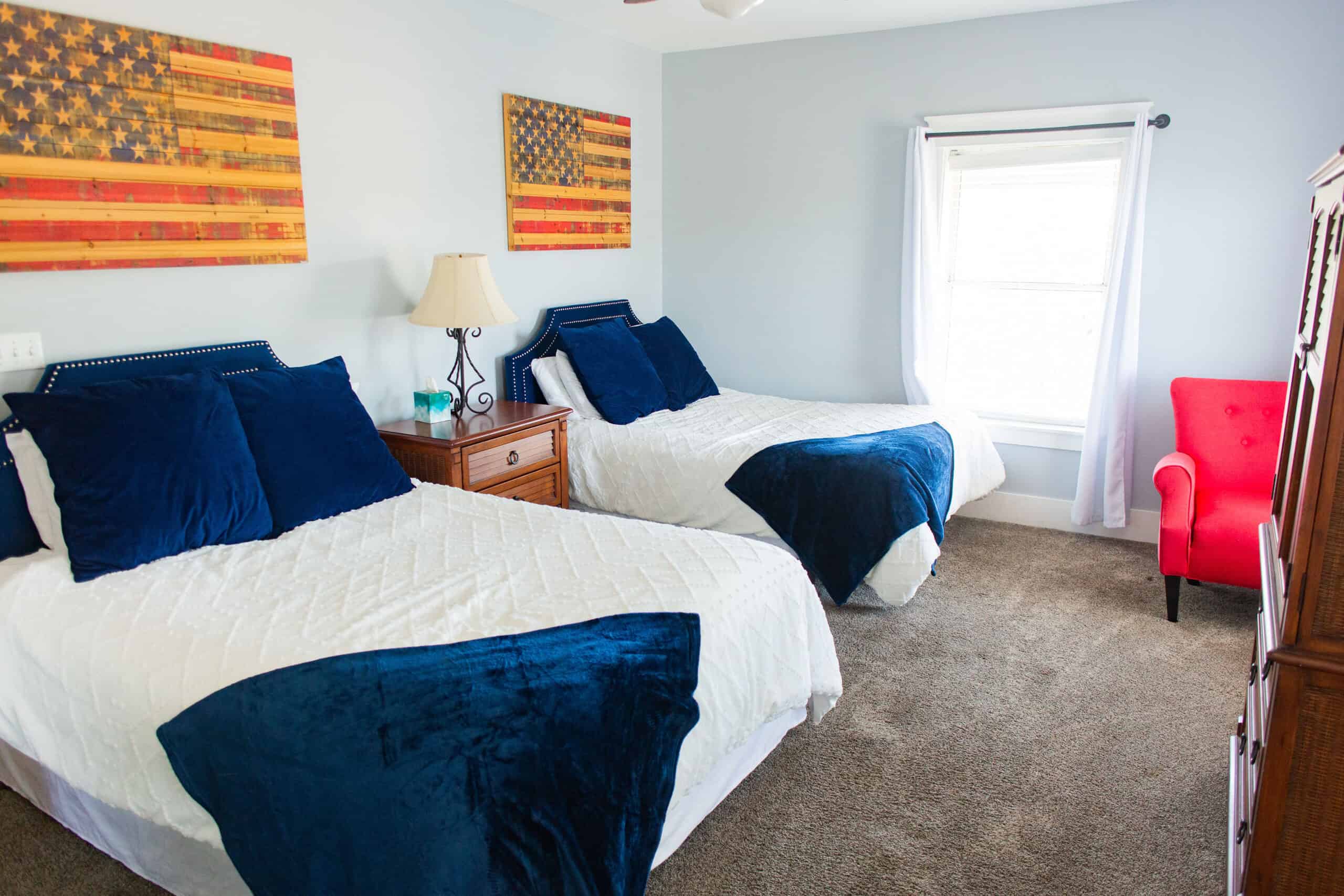 Bedroom with two beds, navy blue bedding, a nightstand, lamp, window with white trim, red chair, and two American flag artworks on the wall.