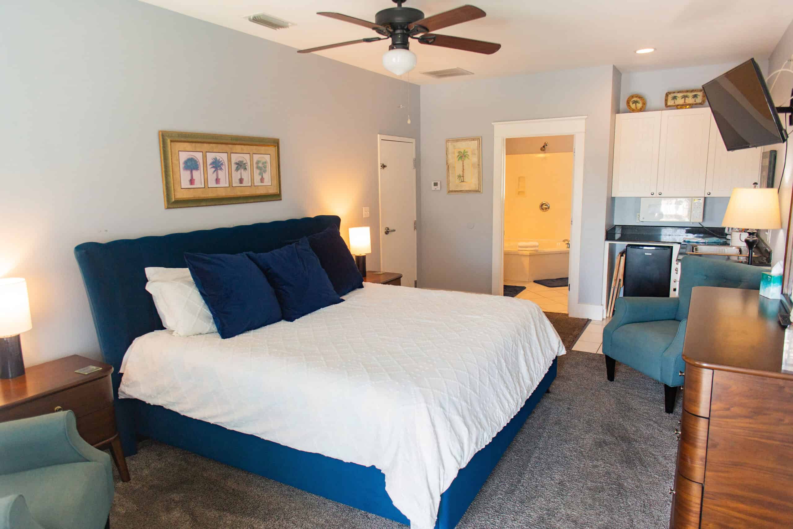 Bedroom with a large bed, blue accents, two chairs, a ceiling fan, lamps, a wall-mounted TV, and a glimpse of a bathroom in the background.