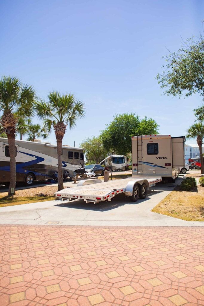 A travel trailer with an empty flatbed trailer is parked on a paved area near palm trees, with other RVs and greenery in the background on a sunny day.