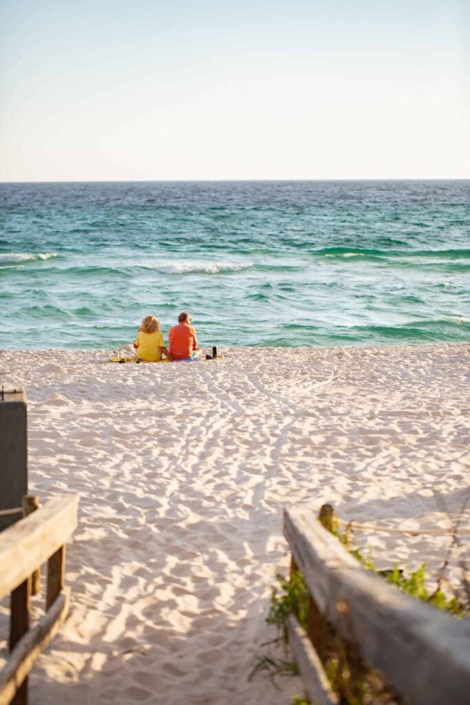 Two people sit on the sandy beach facing the ocean, with waves in the background and wooden railings framing the path to the shore.