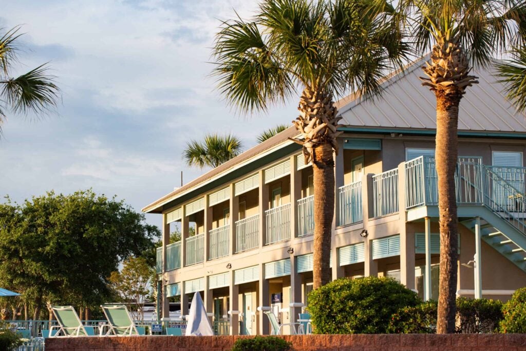 Two-story beach resort building with blue railings, surrounded by palm trees and lounge chairs near a pool area, under a partly cloudy sky.