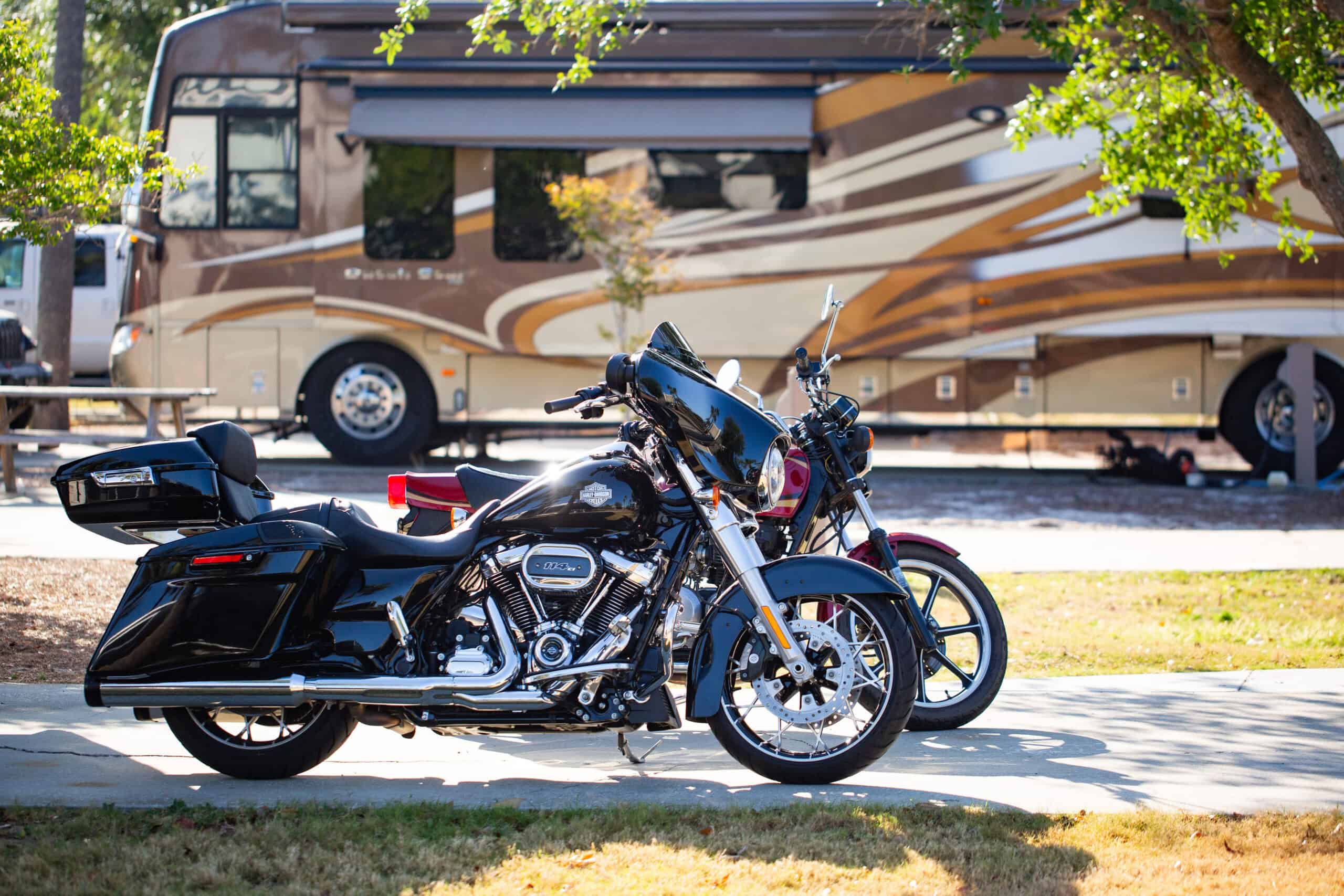 Two parked motorcycles in front of a beige and brown RV on a sunny day, with trees and grass visible in the background.