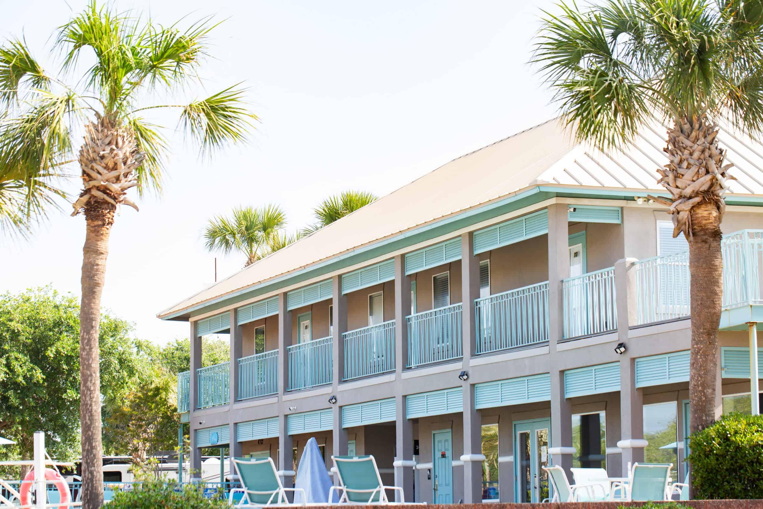 Two-story motel with light blue railings, palm trees, and patio chairs arranged near the front under a clear sky.