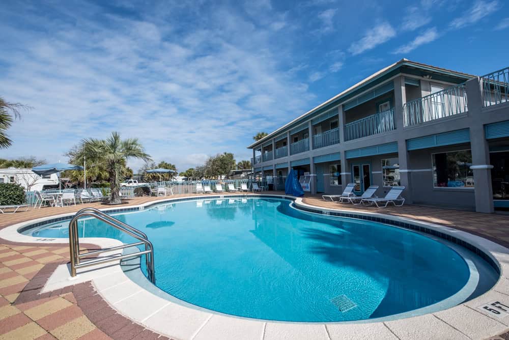 Outdoor swimming pool surrounded by lounge chairs and a two-story hotel building under a partly cloudy sky.