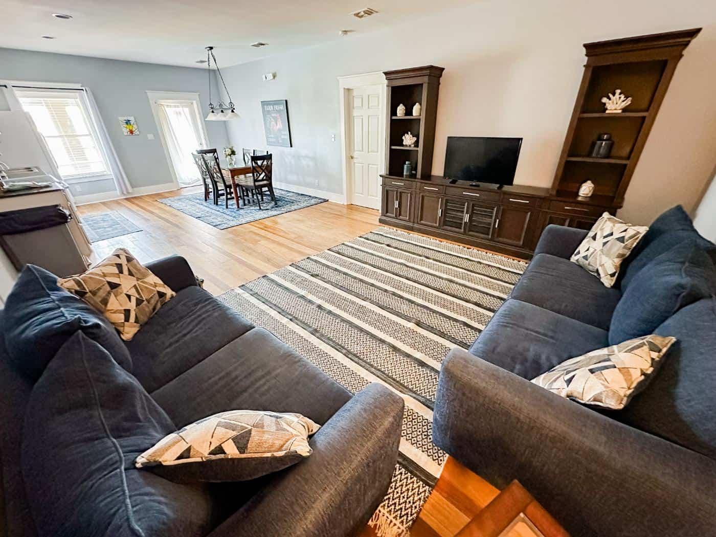 Spacious living room with two blue sofas, a striped rug, TV on a wooden stand, and a dining area in the background.